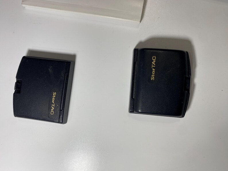 Puede incluir: Dos bater&iacute;as rectangulares negras para tel&eacute;fono m&oacute;vil de un dispositivo StarTAC. Cada unidad presenta un acabado mate liso con el nombre de la marca StarTAC impreso en letras doradas en la parte frontal. Los componentes est&aacute;n colocados lado a lado.