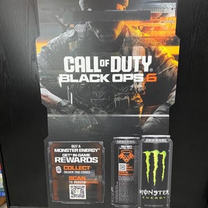 May include: A promotional cardboard standee for Call of Duty Black Ops 6 featuring a soldier in a beanie. The display includes text about Monster Energy in-game rewards, a QR code, and images of two black Monster Energy cans with green logos.