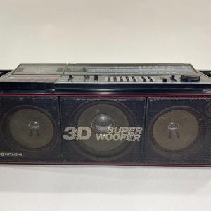 May include: A black Hitachi portable stereo boombox featuring a cassette deck, radio tuner, and equalizer sliders on the top panel. The front displays three speaker grilles with red trim, and the text 3D SUPER WOOFER printed in white.