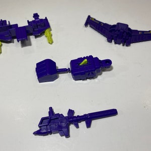 May include: Several disassembled purple and green plastic toy parts are scattered on a white surface. The parts include a wing-like structure, a rectangular piece with green accents, and a long, gun-shaped component. The pieces appear to be from a larger toy.