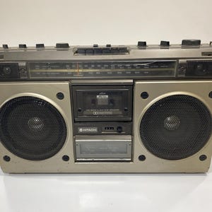 May include: A vintage Hitachi portable stereo boombox in a metallic silver finish. It features two large circular speakers with black mesh grilles, a central cassette deck, a radio tuner display with frequency markings, and various control knobs.