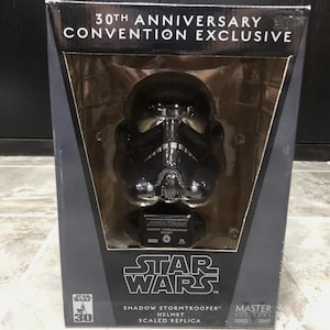 以下が含まれることがあります： 黒い箱に入った、黒と銀のスターウォーズシャドウストームトルーパーヘルメットレプリカ。箱は黒で、白い文字で「30周年記念コンベンション限定」と「スターウォーズシャドウストームトルーパーヘルメット縮尺レプリカ」と書かれています。