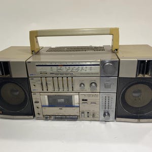 以下が含まれることがあります： シルバーとブラックのヴィンテージJVCブームボックス。このポータブルステレオは、カセットデッキ、ラジオチューナー、スピーカーを備えています。ベージュのキャリーハンドルとレトロなデザインです。