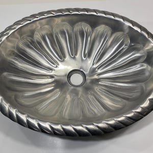 May include: Oval-shaped, silver-colored metal bundt pan with a decorative rope-like border. The interior features a fluted design radiating from a central hole. The pan is made of a shiny, reflective metal.