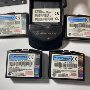 Puede incluir: Una unidad base de tel&eacute;fono m&oacute;vil Motorola negra y cuatro bater&iacute;as de iones de litio rectangulares. Cada bater&iacute;a tiene una etiqueta blanca con el logotipo de Motorola, c&oacute;digo de barras y texto que incluye SNN5651C, SNN4B14B y advertencias.