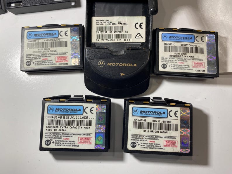 Puede incluir: Una unidad base de tel&eacute;fono m&oacute;vil Motorola negra y cuatro bater&iacute;as de iones de litio rectangulares. Cada bater&iacute;a tiene una etiqueta blanca con el logotipo de Motorola, c&oacute;digo de barras y texto que incluye SNN5651C, SNN4B14B y advertencias.