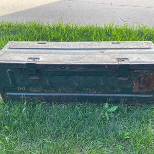 May include: A rectangular olive green metal ammunition box with a hinged lid and two metal latches. The exterior features weathered paint, rust patches, and stenciled text including "TNT" and "LOT" markings across the front and top surfaces.