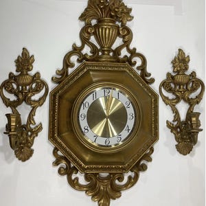 May include: A gold-colored wall clock with an octagonal frame, featuring a white clock face with black numerals. Two matching sconces flank the clock. The set has ornate, floral-inspired detailing, creating a vintage aesthetic.