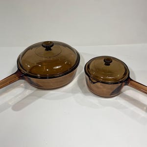 May include: Two amber-coloured glass saucepans with lids. The pans have long handles and are of different sizes. The lids have small, round knobs. The glass has a slightly textured appearance. The pans are on a white surface.