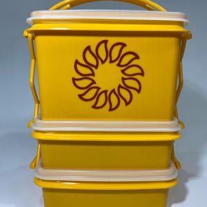 Könnte beinhalten: Ein Stapel aus drei gelben Lebensmittelbehältern mit cremefarbenen Deckeln und einem gelben Griff. Der obere Behälter hat ein weinrotes Sonnenblumen-Design. Die Behälter sind aus Kunststoff.