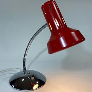 May include: A red desk lamp with a chrome base and a flexible, silver-colored neck. The lamp's shade has vertical slits and is angled downward. The base is round and reflective, with a small on/off switch. The lamp is plugged into a wall outlet.