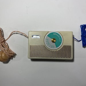 May include: A rectangular off-white transistor radio with a gold-toned metal speaker grille. The device features a circular dial with a teal accent and the word BROADCAST printed in white. A tan cord with a plug and a blue wire are attached.