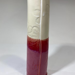 May include: A tall, handmade ceramic vase with a two-tone design. The top half is a creamy white with embossed floral patterns, while the bottom half is a deep burgundy. The vase has a rectangular shape with slightly rounded edges.
