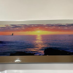 May include: A rectangular wall art piece featuring a sunset over the ocean with a lighthouse in the distance. The image is framed by a wide, reflective mirrored border with four silver-toned circular mounting studs at each corner of the frame.