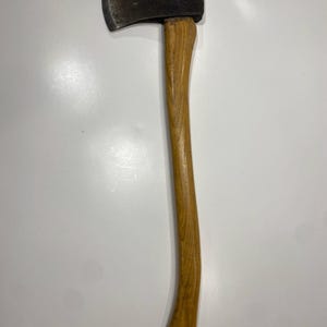 May include: A single-bit axe featuring a dark, weathered metal head with visible signs of use and a smooth, light brown wooden handle with a curved grip. The tool has a sharp, slightly worn cutting edge and a sturdy, elongated wooden shaft.