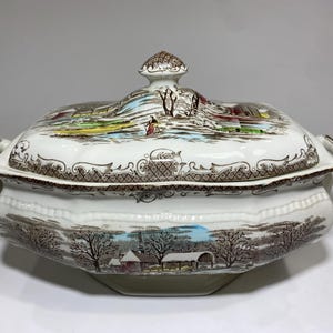 May include: A lidded serving dish with a landscape design in brown, blue, and green. The white base has a decorative rim and handles. The lid features a knob and a detailed landscape scene. The dish is likely made of ceramic.