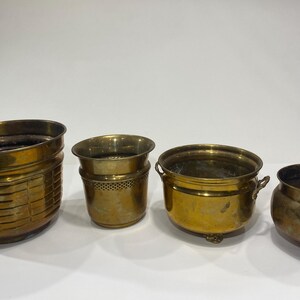 May include: A set of four brass planters of varying sizes and shapes. The first features a horizontal grid pattern, the second has a perforated rim, the third includes small side handles and feet, and the fourth is a smooth, rounded bowl with feet.
