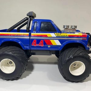 May include: A blue toy truck with large black tires and white rims. The truck features a black roll bar, chrome engine details on the hood, and red, yellow, and white side decals. The text "4x4" and "OFF ROADER" are printed on the side panels.