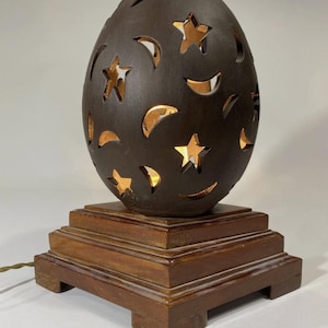 May include: A dark brown egg-shaped lamp with star and crescent cutouts. The lamp sits on a square wooden base and has a power cord. The cutouts allow light to shine through, creating a warm glow. A decorative lighting fixture.