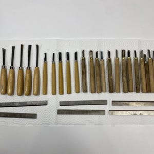 May include: A collection of wood carving tools with light-colored wooden handles and metal tips, arranged in a row. Below the tools are six rectangular metal sharpening stones of varying lengths, all laid out on a white textured surface.