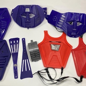 May include: A collection of plastic toy armor pieces including two purple hexagonal chest plates, two purple face masks, two red chest plates with metallic decals, a silver curved arm guard, and two purple plastic straps with slotted cutouts.
