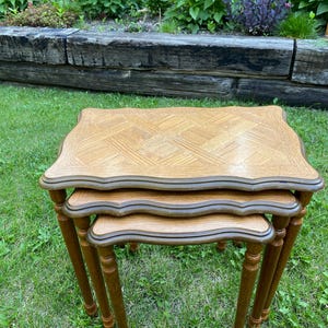 May include: A set of three nested tables featuring light brown wood tops with a herringbone pattern and scalloped edges. Each table has four slender, turned wooden legs in a matching medium brown finish, designed to stack neatly together.