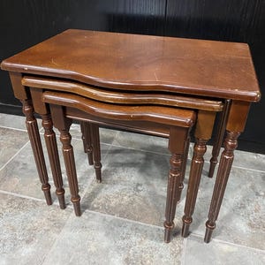May include: Set of three antique nesting tables. The tables are made of dark brown wood with a glossy finish. Each table has a rectangular top with a curved edge and decorative, turned legs. The largest table is approximately 61 cm wide.