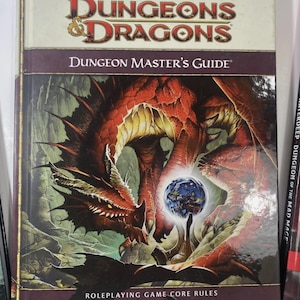 May include: A hardback book titled "Dungeons & Dragons Dungeon Master's Guide." The cover shows a red dragon with a blue orb. The book is a roleplaying game core rule book by James Wyatt.