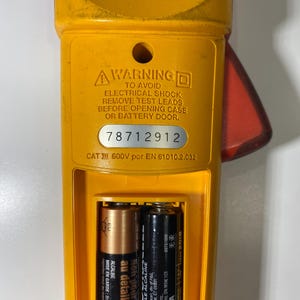 Pu&ograve; includere: Un multimetro a pinza giallo e rosso con vano batteria aperto contenente due batterie AA. Il dispositivo presenta un'etichetta di avvertenza sulle scosse elettriche, il numero di serie 78712912 e la classificazione CAT III 600V EN 61010.2.032.