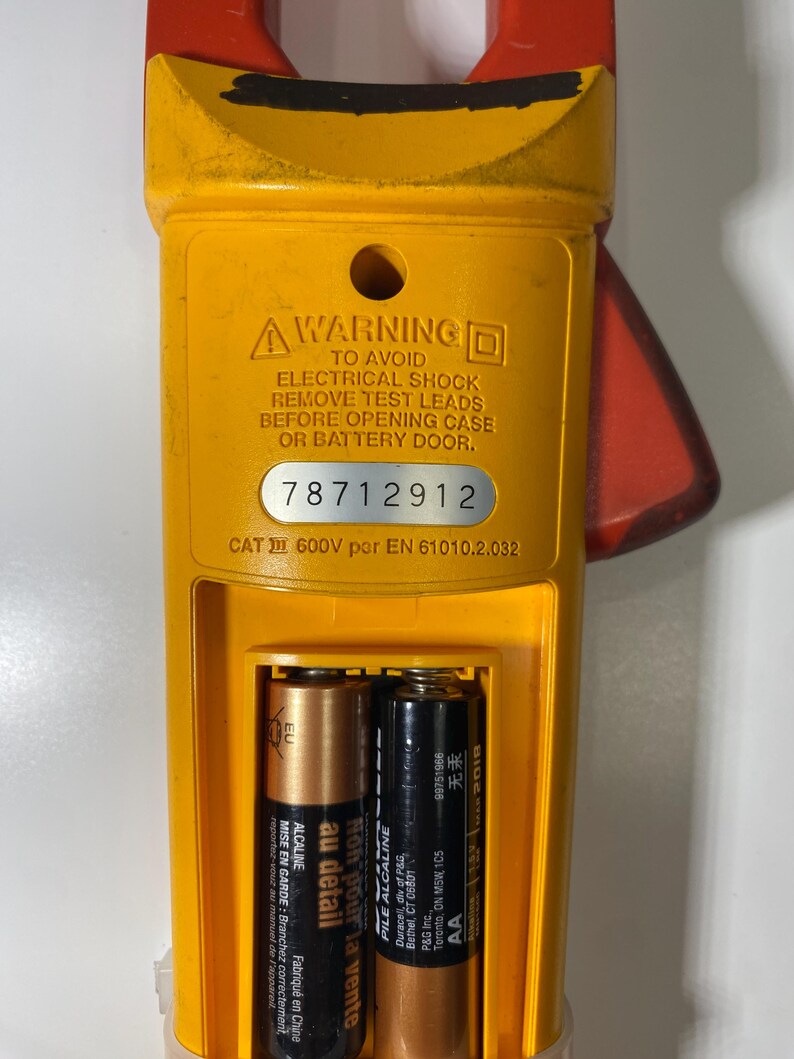 Pu&ograve; includere: Un multimetro a pinza giallo e rosso con vano batteria aperto contenente due batterie AA. Il dispositivo presenta un'etichetta di avvertenza sulle scosse elettriche, il numero di serie 78712912 e la classificazione CAT III 600V EN 61010.2.032.