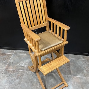 May include: A wooden rocking chair with a light brown finish. The chair has a high back with vertical slats, armrests, and a footrest. The seat cushion is a gold color. The chair is designed for a child.