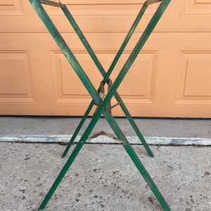 May include: A green metal folding stand with an X-shaped frame design. The structure features four long legs that cross at a central pivot point, supporting a rectangular top frame. The metal surface shows signs of wear and weathered paint throughout.