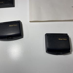 Puede incluir: Dos tapas de bater&iacute;a de tel&eacute;fono m&oacute;vil Motorola StarTAC negras con letras doradas, acompa&ntilde;adas de una vista parcial de una tercera tapa y un manual blanco con el texto RAZR V3c CDMA y motorola.com impreso en tinta negra.