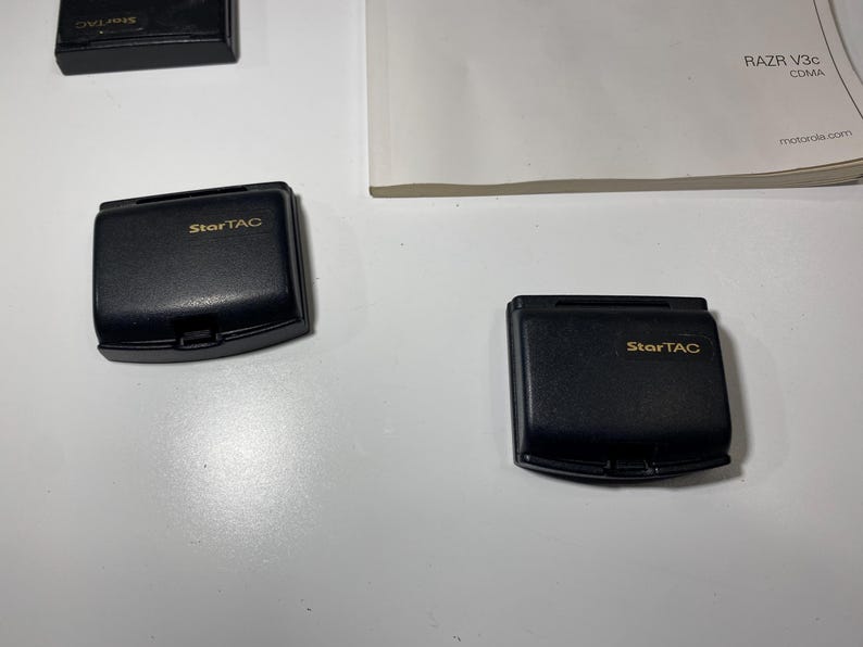 Puede incluir: Dos tapas de bater&iacute;a de tel&eacute;fono m&oacute;vil Motorola StarTAC negras con letras doradas, acompa&ntilde;adas de una vista parcial de una tercera tapa y un manual blanco con el texto RAZR V3c CDMA y motorola.com impreso en tinta negra.