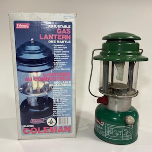 May include: A green Coleman adjustable gas lantern with a clear glass globe and a metal handle. The lantern is next to its original box, which features the Coleman logo and product information. The box reads "Adjustable Gas Lantern, One Mantle."