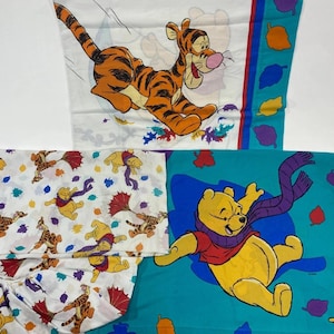 May include: A collection of Winnie the Pooh themed bedsheets. One sheet features Tigger, another has Pooh Bear, and the others have a pattern of Pooh and Tigger with autumn leaves. The sheets are white, teal, and patterned.