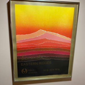 May include: Framed art print depicting a layered mountain landscape in orange, red, and brown, with a yellow sky. The print includes the text "Eastern Ridges" and the date "31/5 - 15/6/1979". The artwork is encased in a gold-coloured frame.