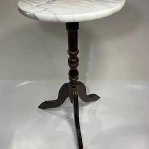 May include: A round side table featuring a white marble top with grey veining. The table is supported by a dark brown wooden pedestal base that transitions into three curved legs, providing a stable foundation for the circular stone surface.