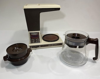 May include: A vintage General Electric coffee maker in cream and dark brown. The unit features a tall tower, a warming plate, and a control panel with buttons. Included are a separate dark brown plastic filter basket and a clear glass carafe.
