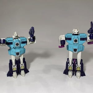 May include: Two blue and white Transformers action figures with purple accents. The figures are posed in a standing position with their arms outstretched.