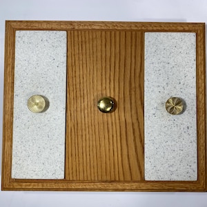 Peut inclure: Un plateau rectangulaire en bois avec une finition en grain de chêne naturel. Le plateau comprend deux panneaux verticaux en pierre blanche mouchetée sur les côtés gauche et droit, et un panneau central en bois. Trois boutons en laiton sont fixés.