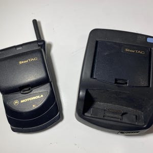 Puede incluir: Un tel&eacute;fono m&oacute;vil plegable Motorola StarTAC de color negro con una peque&ntilde;a antena externa y una base de carga negra a juego. Ambos dispositivos presentan el logotipo StarTAC en dorado y el tel&eacute;fono muestra la marca Motorola en letras doradas.