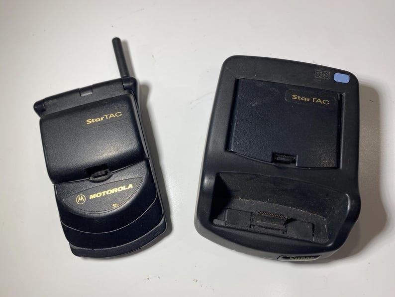 Puede incluir: Un tel&eacute;fono m&oacute;vil plegable Motorola StarTAC de color negro con una peque&ntilde;a antena externa y una base de carga negra a juego. Ambos dispositivos presentan el logotipo StarTAC en dorado y el tel&eacute;fono muestra la marca Motorola en letras doradas.