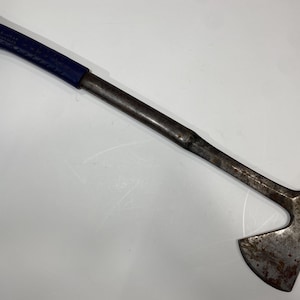 May include: A metal hatchet with a long, straight handle featuring a dark blue textured grip. The head of the tool is made of weathered, dark grey steel with visible signs of oxidation and wear along the blade edge and the flat poll surface.