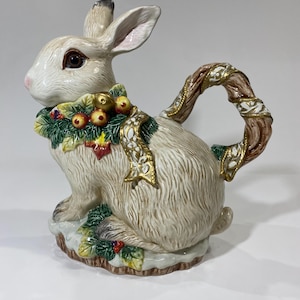 May include: A ceramic teapot in the shape of a rabbit. The rabbit is off-white with a decorative green and gold garland around its neck. The handle is a brown, twig-like design with gold accents. The teapot sits on a base with green leaves and red berries.