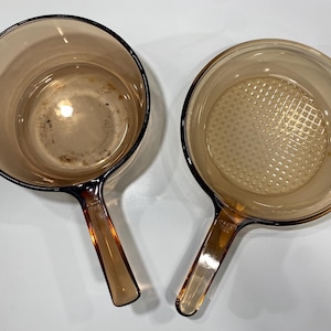 May include: Two amber-coloured glass saucepans with long handles. The left saucepan has a smooth interior base, while the right saucepan features a textured grid pattern on the interior bottom. Both pieces are made of translucent brown glass.