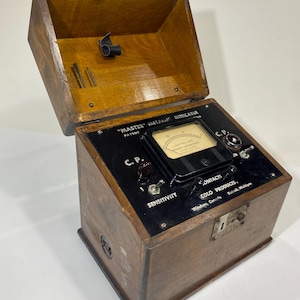 May include: A vintage Master Metallic Indicator device housed in a polished wooden box with a hinged lid. The black control panel features a central analog gauge, two toggle switches, two knobs, and text reading Master Metallic Indicator, C.P., and Colo Products.