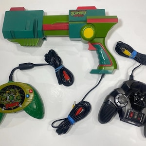 May include: A green and red Teenage Mutant Ninja Turtles toy blaster, a green oval-shaped controller with a TMNT graphic, and a black Darth Vader-themed video game controller. Each device includes a black wired cable with red and yellow RCA plugs.