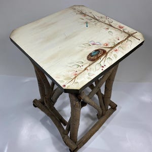 May include: A square side table with a cream-colored top featuring a hand-painted design of flowering branches, leaves, and a bird's nest with blue eggs. The base is constructed from four thick, textured brown tree branches arranged vertically.