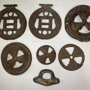May include: A collection of seven dark brown, weathered metal hardware pieces. Two circular items feature a central H-shaped cutout, one has an ornate swirling pattern, three are circular discs with four-spoke cutouts, and one is a small handle.